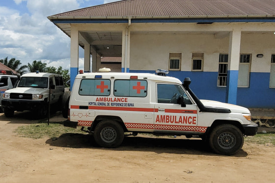 A white 4x4 with red lettering reading AMBULANCE parked on a dirt road outside a one-story concrete building.