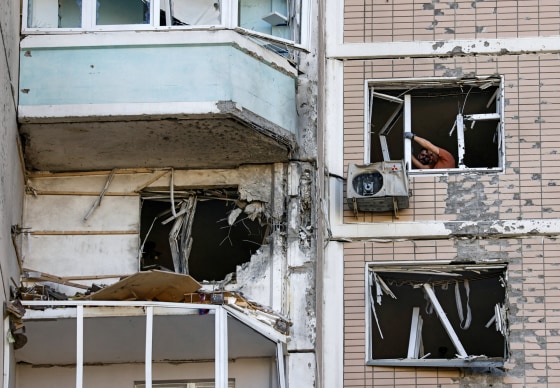 Residents clear broken windows in a damaged apartment building, as seen from the outside of the building