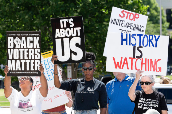People hold signs that say "Black Voter Matter", "It's About Us", and "History Is Watching" while standing outside