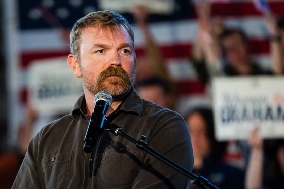 Graham Platner stands behind a microphone near his chin, people are seated behind him holding signs