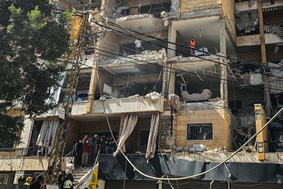 People stand inside of the rooms of an apartment building with the exterior walls blown off