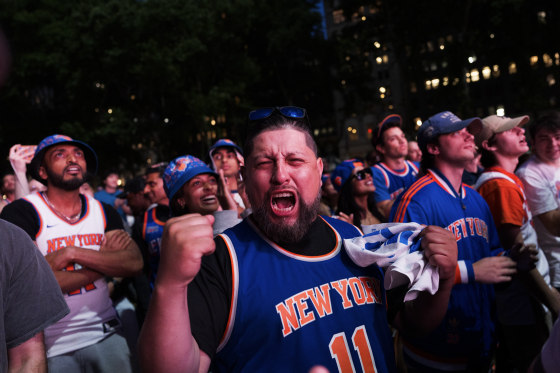A Knicks fan pumps his fist and yells toward the camera while surrounded by fellow fans.