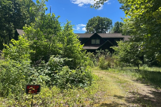 A house is shrouded by trees in a forested area, a sign on the front of the property next to the path leading to the home reads "KEEP OUT"