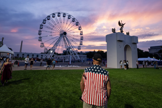 A man in a shirt with stars and stripes of the American flag stands in front of a Ferris Wheel at sunset.