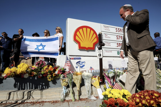 Flowers arranged in front of a gas station sign in a makeshift memorial, with people holding an Israeli flag behind it.