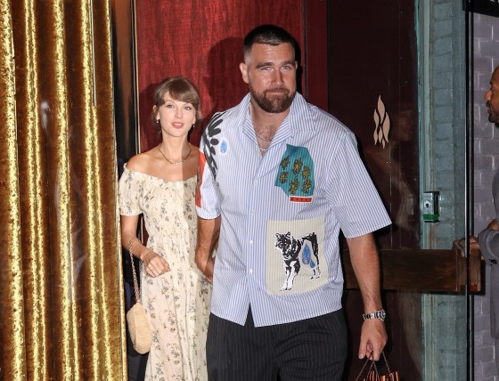 Travis Kelce, looking toward the camera, holds Taylor Swift's hand as they exit a building to the street.