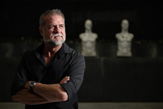 Victor Marx stands for a portrait with his arms crossed, two busts are seen at a shooting range in the distance behind him