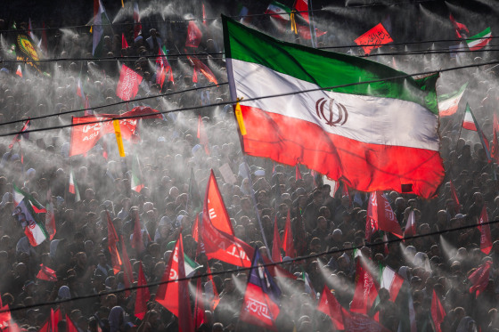 A large crowd of people on the street, holding flags, including a large Iranian flag