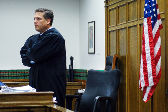 Roger Rogoff stands inside of a courtroom, an American flag hangs off of the wall