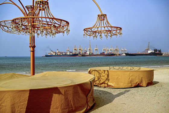 Two tables with chandeliers on a beach in front of large container ships floating in the distance.