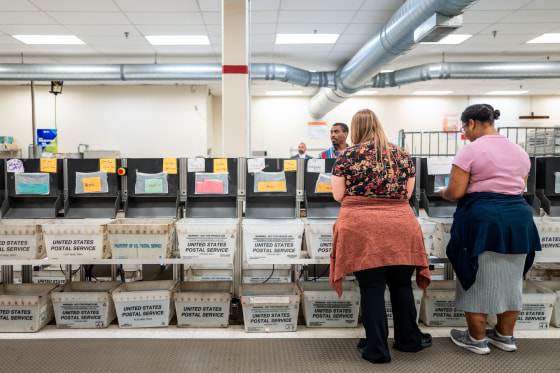 Two people stand in front of a row of United States Postal Service bins