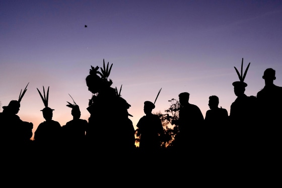 Indigenous people wearing large bird feathers on their heads silhouetted against a dark sky at sunrise.