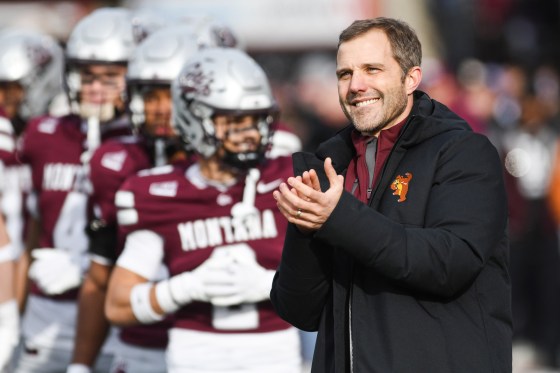 Seth Bodnar stands and claps while smiling near blurred football players outside on the field