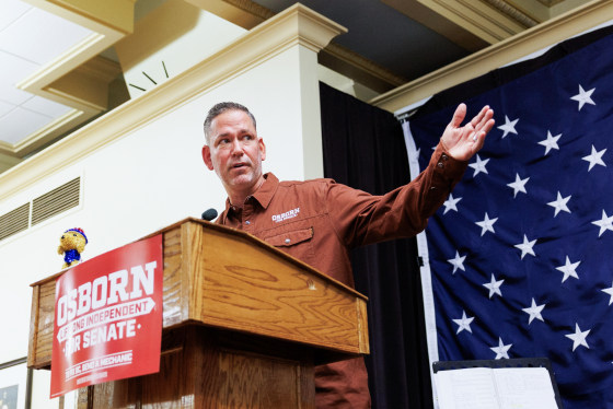 Dan Osborn speaks at a podium and extends his hand outwards, there is a red poster on the front of the podium that reads in white text: "Osborn, lifelong independent, for senate. To fix DC, send a mechanic."