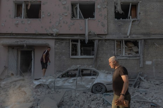 A man walks past while a man stands on a destroyed car  following an air attack.