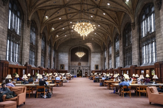 A large stone room with a vaulted ceiling and chandelier and long rows of wooden desks.