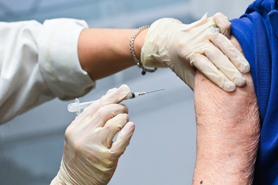 A gloved hand moves a syringe towards an arm with wrinkled skin, while holding the shoulder with their other gloved hand