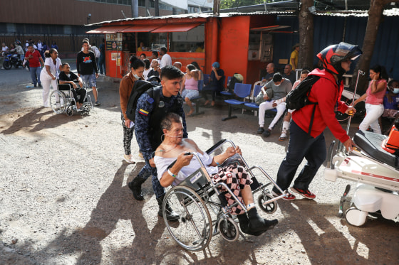 People in wheelchairs are assisted outside, while others sit in waiting room chairs that have been placed outside