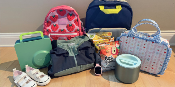 Back-to-school gear for kids laid out on a wood floor: two backpacks (pink strawberry print and navy), a green lunchbox, white toddler sneakers, a folded jacket, Little Spoon snack pouches, a kids' smartwatch, an insulated food container, and a floral gingham lunch bag.