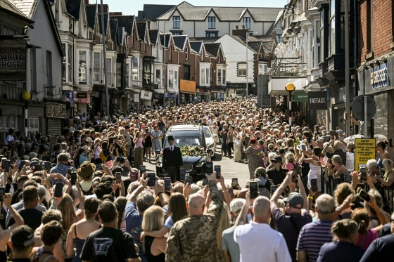 Hundreds of people crowding a street around a black hearse.