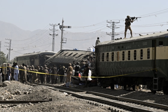 A crowd of people and soldiers gathered near an overturned and smashed train car on the tracks.
