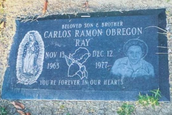 Carlos Ramon Obregon's headstone, flat on the ground, showing an engraving of Mother Mary on the left, a praying angel in the center, and Carlos on the right. The full headstone reads "Beloved Son & Brother, Carlos Ramon Obregon, "Ray", Nov. 13, 1963 - Dec. 12, 1977, You're forever in our hearts"