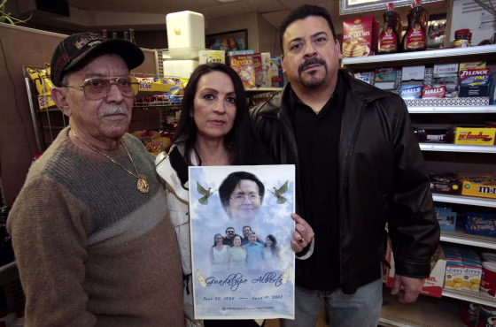 Image: Abraham Alberto stands in his grocery store with his children holding a memorial poster showing his deceased wife who was killed in 2008  when the 2005 Toyota Camry she was driving crashed in Flint