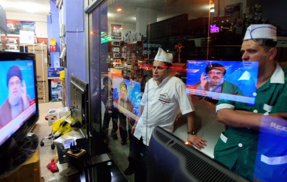 Workers at a nearby restaurant watch the televised speech of Hezbollah leader Hassan Nasrallah as they stand outside a shop in the southern port city of Sidon, Lebanon, on June 24.