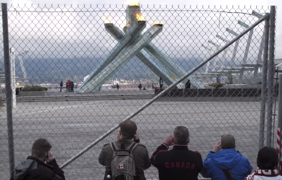 Image: People take pictures of the Olympic cauldron from behind a fence keeping the public out of the plaza it is situated in at the Vancouver 2010 Winter Olympics