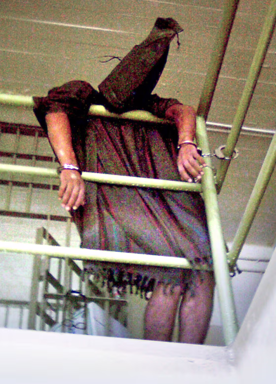 In this undated photo, a hooded Iraqi detainee appears to be cuffed at both wrists and collapsed over a rail at Abu Ghraib prison.