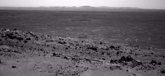 A picture from the Spirit rover's navigational camera shows terrain leading up to the summit of Husband Hill in the foreground, with what appears to be a crater on the plain below and distant hills on the horizon.