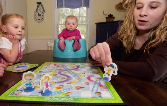 Michelle Hastings, right, plays Chutes and Ladders with her daughter Peyton Hastings, 2, left, as her daughter Andersen, 5 months, center, watches at their home in Holliston, Mass.