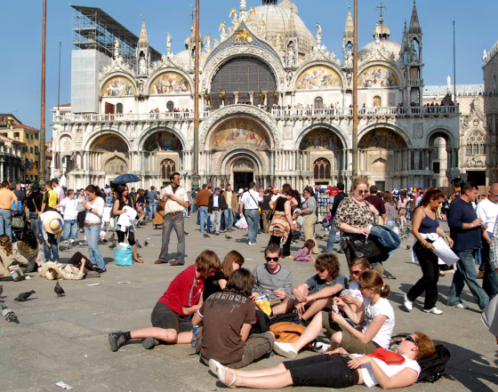 Tourists enjoy the sun in a crowded St. Mark's Square in Venice, northern Italy, with St. Mark's Basilica in the background. A message to visitors to Venice: No bare torsos in St. Mark's Square. No lounging on the monuments. And no feet dangling in the canals.