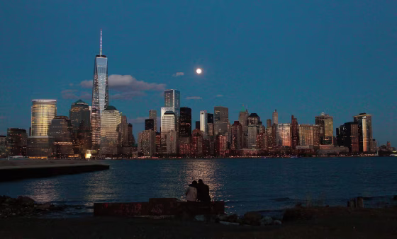 Image: The moon rises behind the skyline of New York's Lower Manhattan as two people watch from a park along the Hudson River in New Jersey