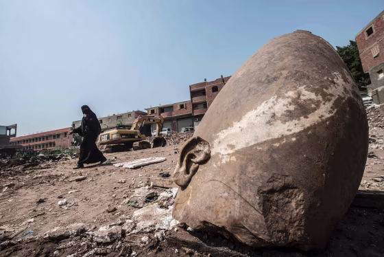Image: An Egyptian woman walks past the head of a statue at the site of a new discovery by a team of German-Egyptian archeologists in Cairo's Mattarya district