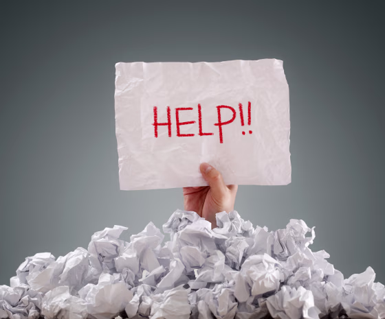 Image: Businessman buried under crumpled pile of papers holds up a help sign