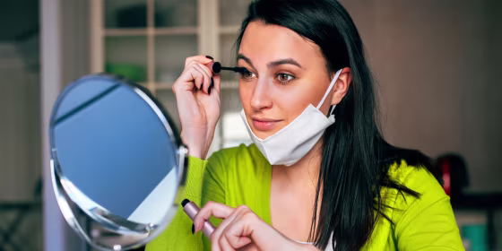 Woman wearing a mask and applying mascara. Scott Barnes and more celebrity makeup artists recommend the best long-lasting makeup to wear while wearing a face mask during Coronavirus 2020.
