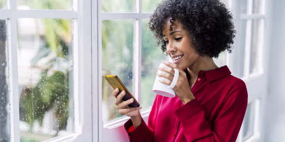 Smiling woman with cup of coffee and cell phone standing at the window at home