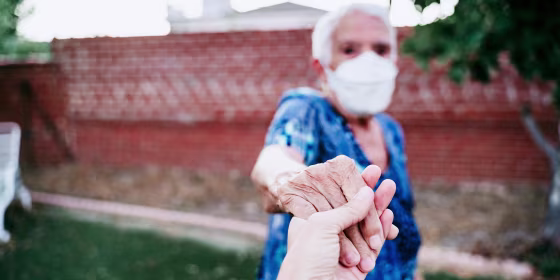 Older woman in backyard wearing an Kn95 mask and holding someones hand. The CDC updated their guidelines on how to shop for face masks and how vaccinated people can socialize. Learn about the updates for face masks and more.