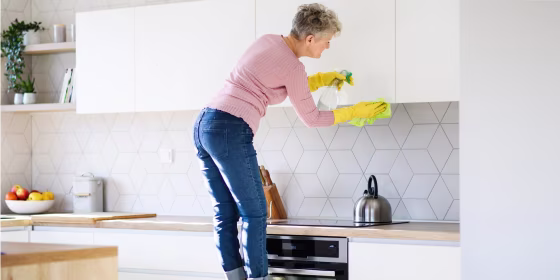 Woman standing on a stool in her kitchen cleaning the outside of her white kitchen cabinets