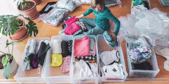 Woman sitting on her floor surrounded by clothes, organizing drawers and big storage bags for under her bed