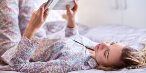 Woman laying in her bed reading a book, wearing a pink matching pajama set