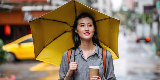 Woman walking outside with a cup of coffee, holding a yellow umbrella