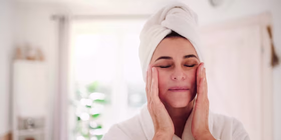 Woman in a robe and towel in her hair, rubbing her cheeks with lotion