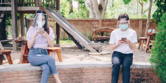 Two Girls 6 feet apart, sitting on a railing at the park wearing face masks and protective eye shields. Shop the best expert-recommended coronavirus eye protection to wear during the pandemic like glasses, goggles and face shields.