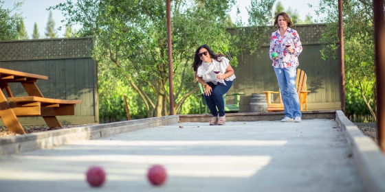 Two woman playing bocce ball outside, while holding wine. Shop the best outdoor games of 2021 to play with family and friends. The best backyard games here for adults and kids include corn hole, outdoor jenga and more.