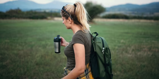 Woman is hiking in nature, with a backpack and holding a travel mug. The best travel mugs of 2021 include reusable cups, travel coffee mugs, ceramic travel mugs and stainless steel travel mugs from Ember, Contigo, YETI and more.