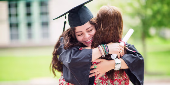 Woman in cap and gown hugging a family member, while holding her diploma