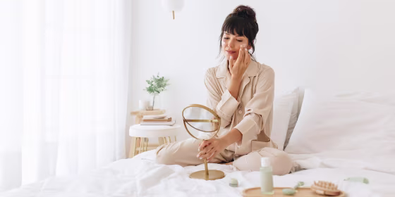 Woman doing self care applying beauty cosmetic, looking into mirror, while sitting in her bed