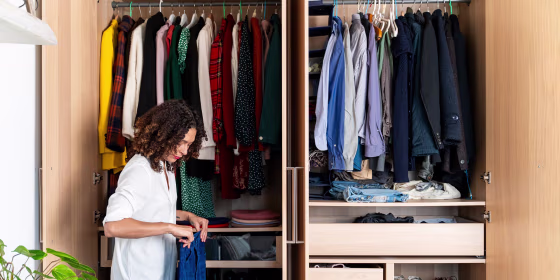 Woman standing next to her closet, holding jeans, clearing out her wardrobe. Here are the best hangers for clothes in 2021. Shop plastic hangers, velvet hangers, and wooden hangers from Amazon, The Container Store and more.